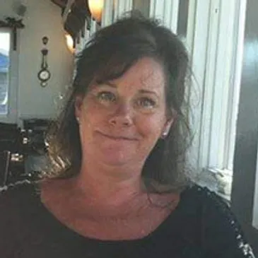 A middle-aged woman with shoulder-length dark hair smiling inside a room with natural light and maritime decor.