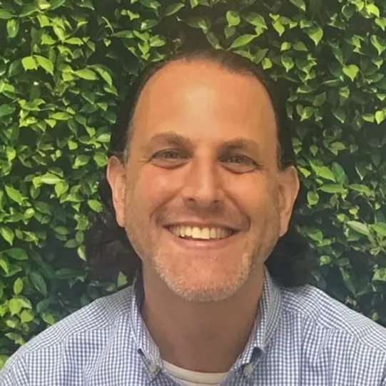 A smiling man with short curly hair, wearing a checkered shirt, poses in front of a leafy green hedge.