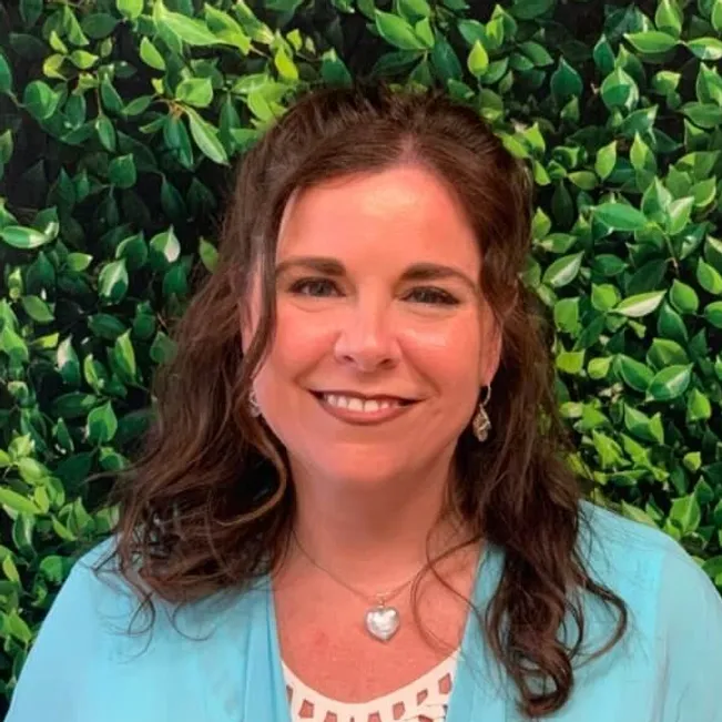 A woman with shoulder-length brown hair smiling against a backdrop of a green leafy wall, wearing a light blue top and heart-shaped necklace.