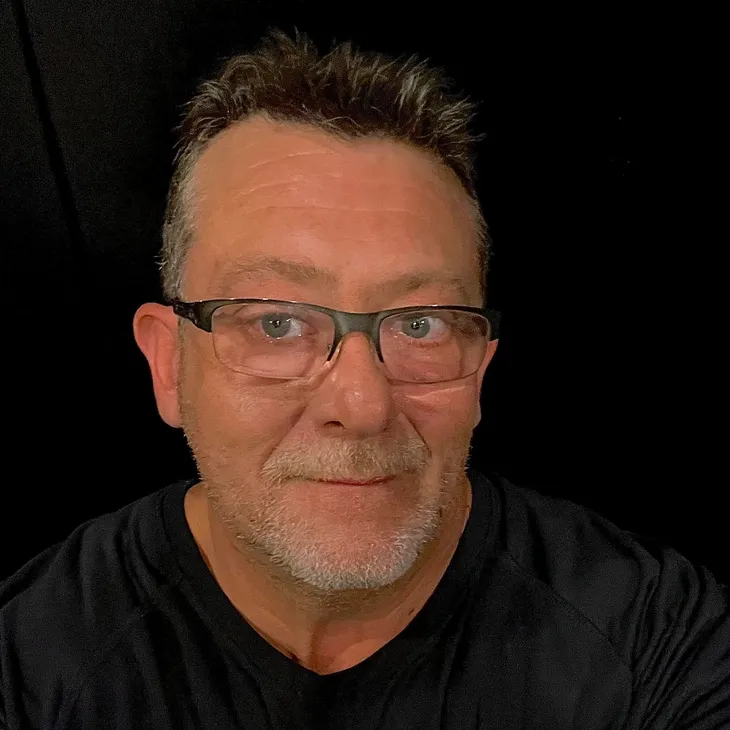 Close-up portrait of a smiling middle-aged man with glasses, featuring short graying hair and a black shirt, against a black background.